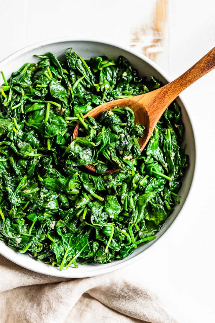 A straight down view of Garlic Sautéed Spinach in a white bowl with a wooden serving spoon on a white background.