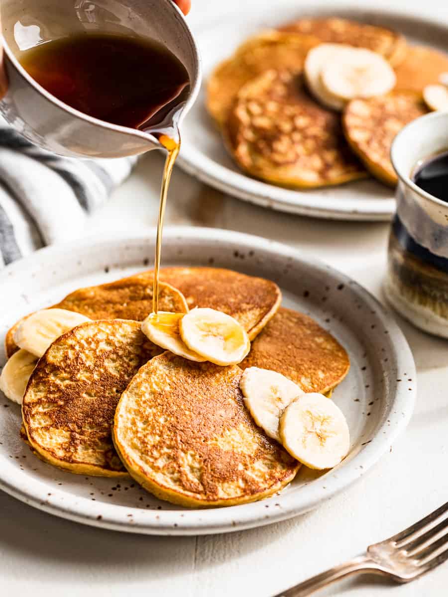 Pouring syrup over a plate of banana pancakes with banana slices over the top.