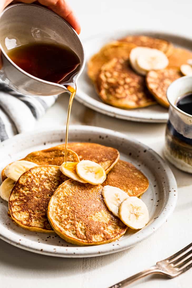 Pouring syrup over a plate of banana pancakes with banana slices over the top.