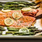 A large sheet pan with Honey Dijon Salmon and Asparagus with a wooden spatula lifting out a piece of salmon.