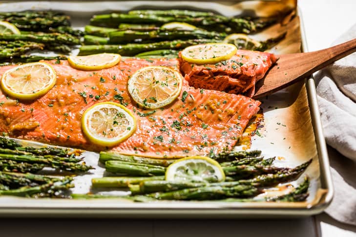 A large sheet pan with Honey Dijon Salmon and Asparagus with a wooden spatula lifting out a piece of salmon.