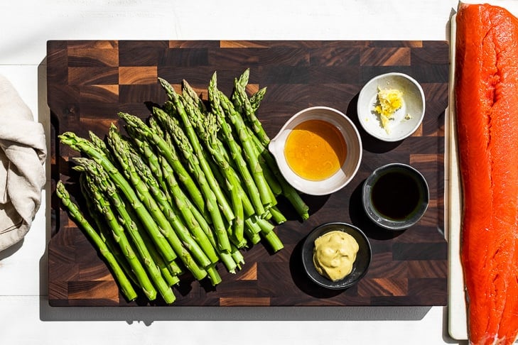 All of the ingredients for this recipe, asparagus, honey, Dijon, garlic, and salmon on a wooden cutting board.