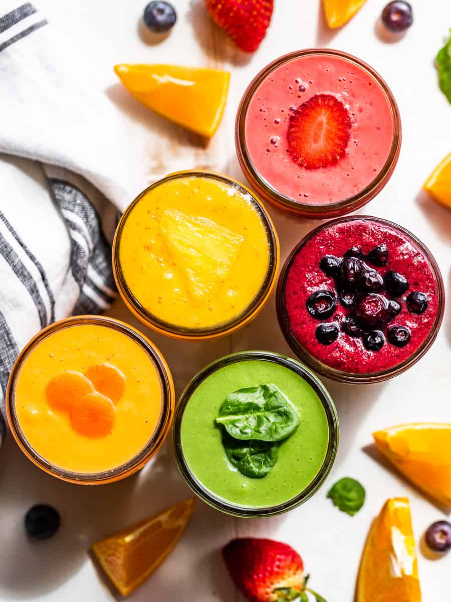 A straight down view of five smoothie flavors, morning glory, mango, pineapple green smoothie, strawberry, and blueberry pie with berries around the smoothies.