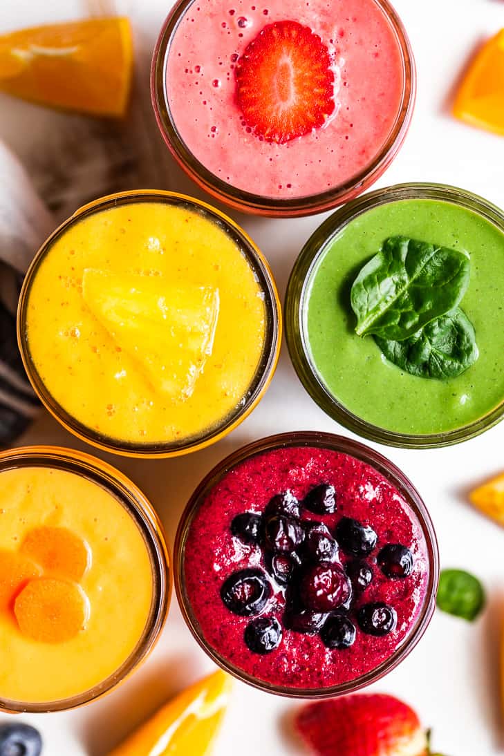 A close up straight down view of five smoothie flavors, morning glory, mango, pineapple green smoothie, strawberry, and blueberry pie with berries around the smoothies.