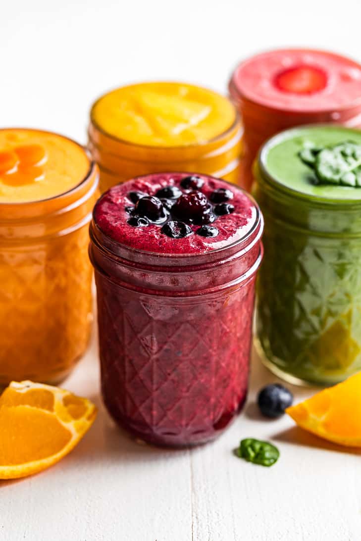 Five smoothies on a white background with orange slices and blueberries around them.