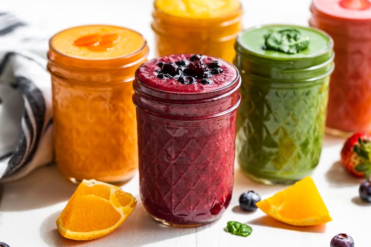 Five smoothies on a white background with orange slices and blueberries around them.