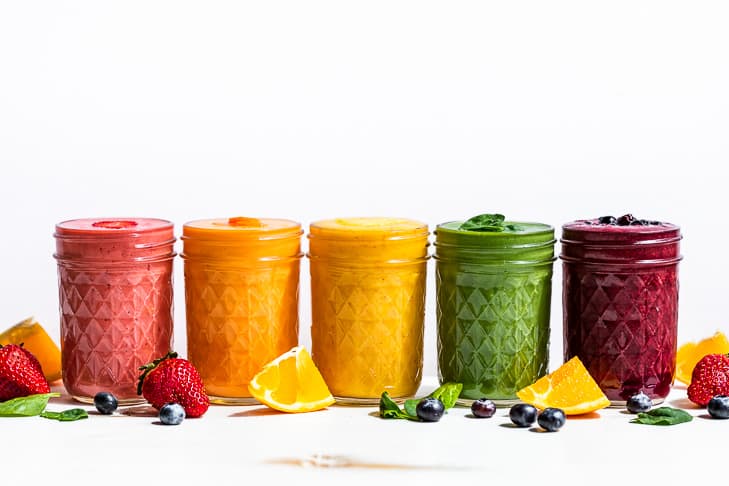 Five smoothies in a row on a white background, strawberry, mango, morning glory, pineapple green smoothie, and blueberry pie smoothie with berries around the smoothies.