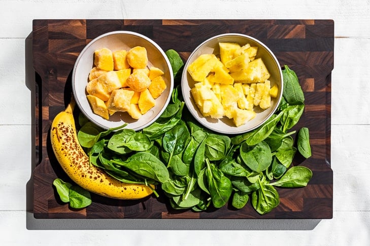 Frozen mango and pineapple, along with a banana and spinach on a wooden cutting board.