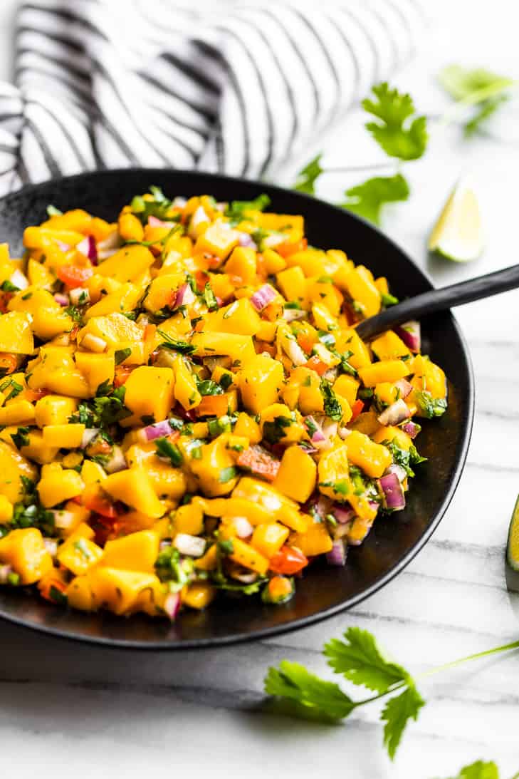 A side view of Mango Salsa in a black bowl with cilantro and lime wedges around it.