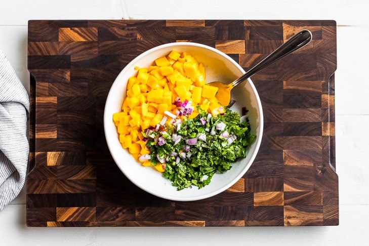 The diced mango, cilantro, onion, and jalapeno in a white bowl on a wooden cutting board.
