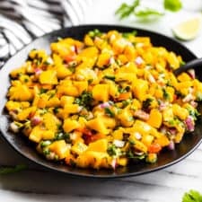 Finished Mango Salsa in a black bowl with a black spoon with lime wedges and cilantro around it.