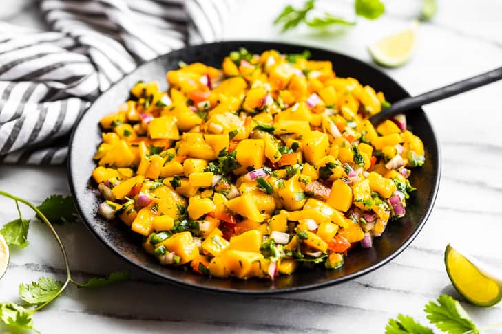 Finished Mango Salsa in a black bowl with a black spoon with lime wedges and cilantro around it.