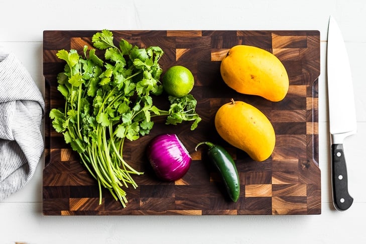 All the ingredients for the mango salsa on a wooden cutting board, mangos, cilantro, jalapeno, onion, and lime.