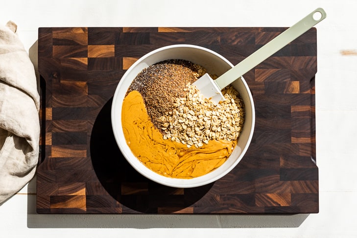 The nut butter, maple syrup, chia and flax seeds, and oats added to a medium sized mixing bowl on a wooden cutting board.