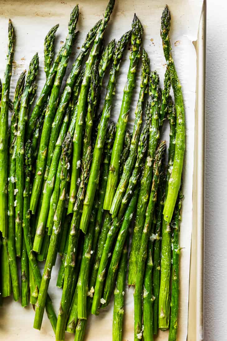 Roasted asparagus on a parchment paper lined gold baking sheet on a white background.