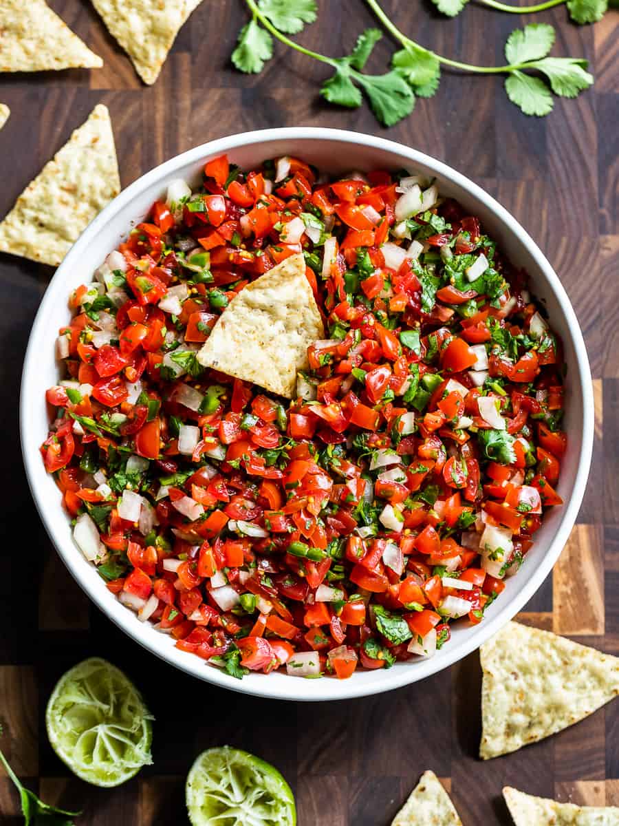 A white bowl of Pico de Gallo on a wooden cutting board with chips around it.
