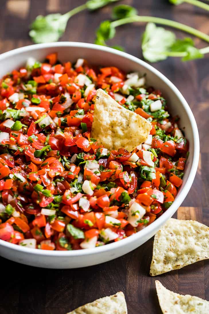 A side view of Pico de Gallo in a white bowl with cilantro and chips around it.