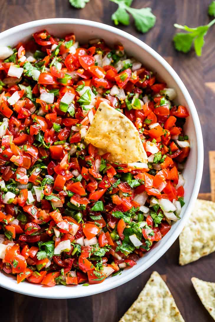 View of half a white bowl filled with Pico De Gallo with a tortilla chip scooping some out.