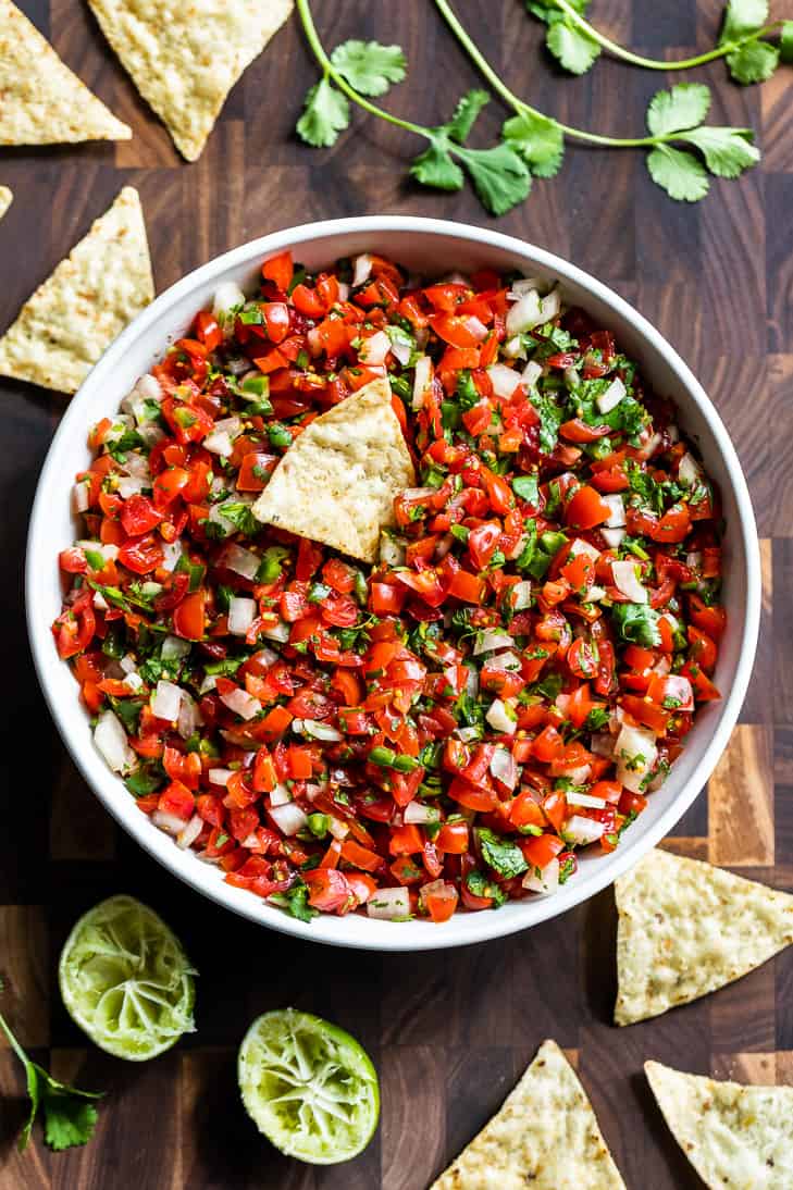 A white bowl of Pico de Gallo on a wooden cutting board with chips around it.