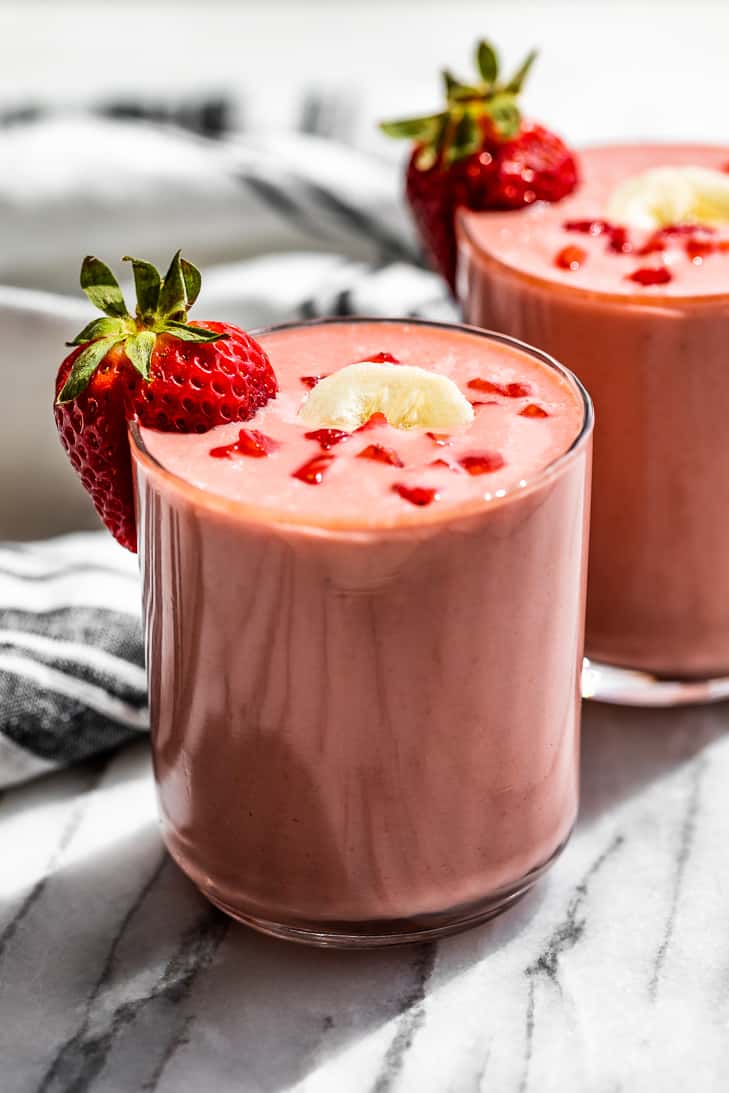 Two glasses Strawberry Banana Smoothie topped with banana sliced and diced strawberries and a blue and white linen striped linen on the side.