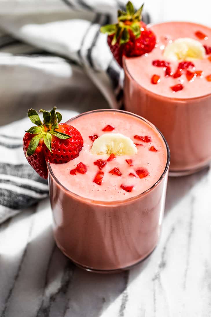 Two glasses Strawberry Banana Smoothie with a blue and white striped linen on the side. The smoothie are topped with banana slices and diced strawberries.