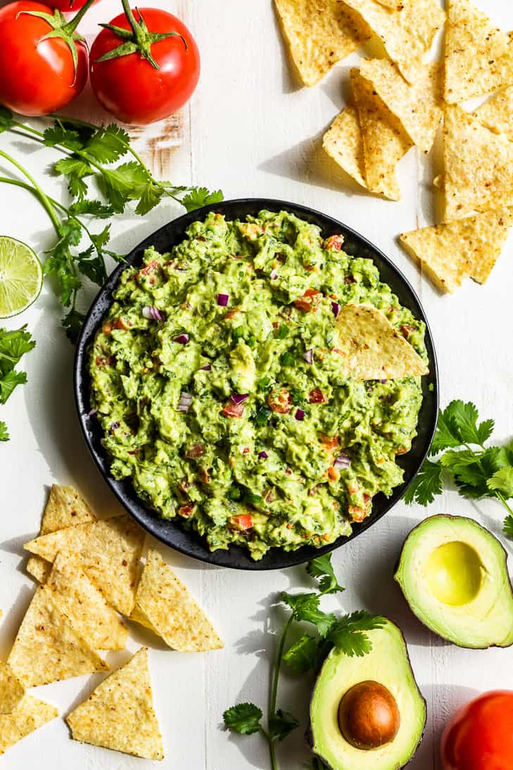 A large bowl of guacamole with chips alongside it and cilantro, tomatoes, and avocados placed around.