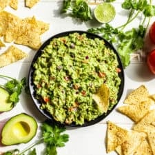 A black bowl of guacamole with chips and cilantro around it.