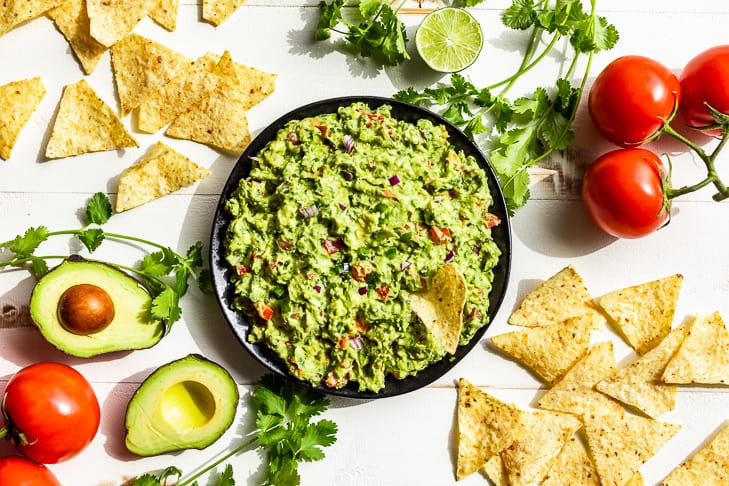 A black bowl of guacamole with chips and cilantro around it.