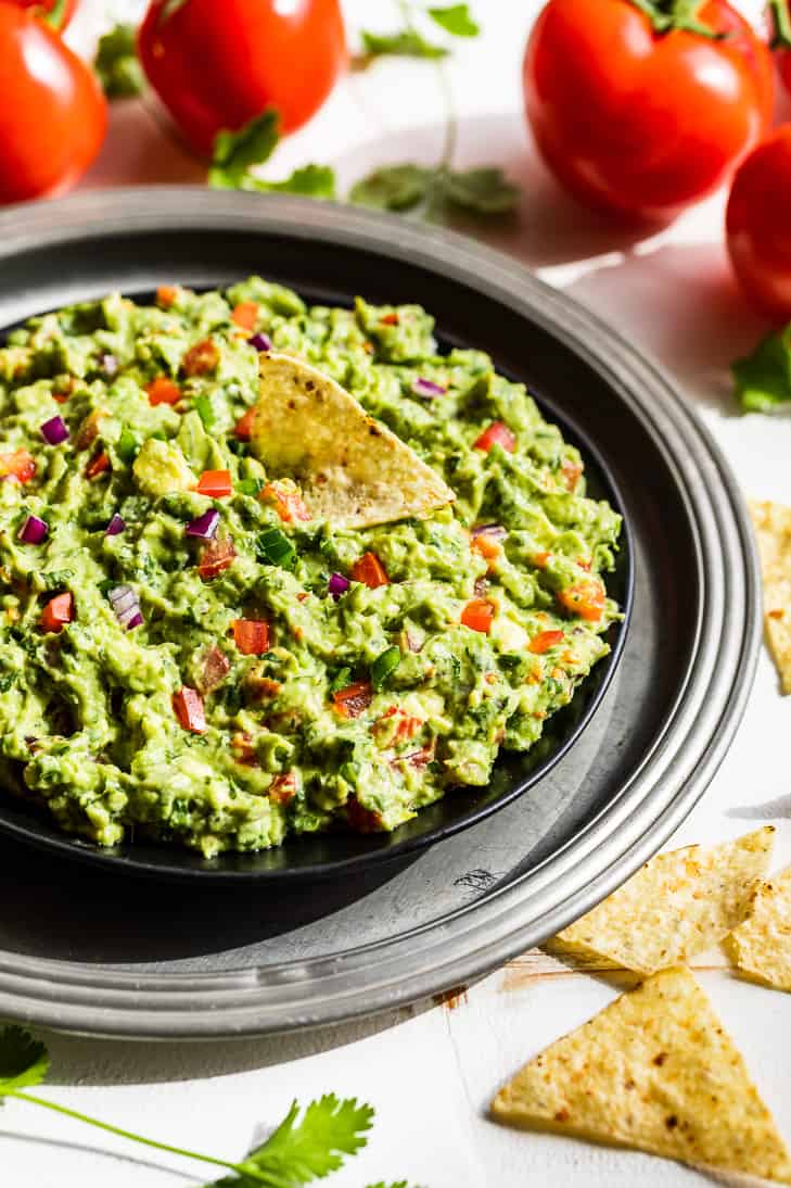 A bowl of guacamole on a silver serving plate with tomatoes an corn chips around it.