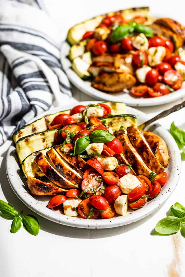 Two pottery plates of Balsamic Grilled Chicken topped with Caprese Salad with a blue and white striped linen on the side with basil sprigs around the plates.