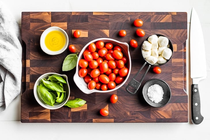 Cherry tomatoes, mozzarella balls, basil, sea salt, and olive oil in pottery bowls on a wooden cutting board.