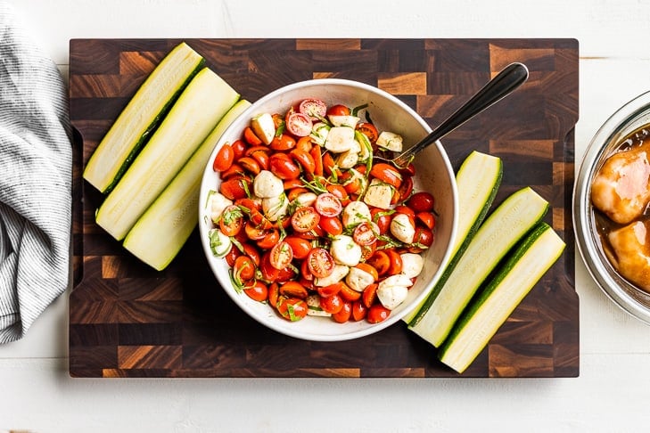 Caprese Salad in a mixing bowl with sliced zucchini on a wooden cutting board.