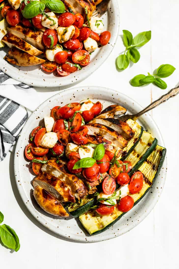 A straight down view of two plates of Balsamic Grilled Chicken topped with Caprese Salad on a white background with basil sprigs around it.