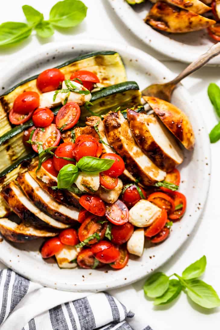 Balsamic Grilled Chicken topped with Caprese Salad on a pottery plate with basil sprigs around it and a blue and white striped linen next to the plate.