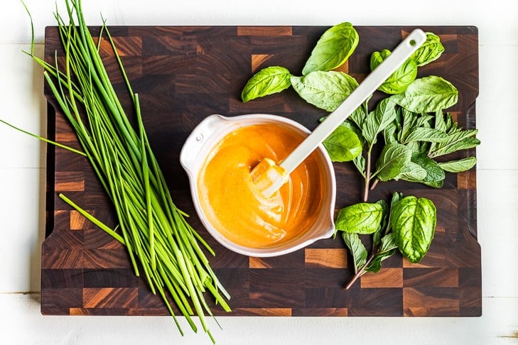 Mixing sriracha mayo in a small bowl on a wooden cutting board with herbs around it.