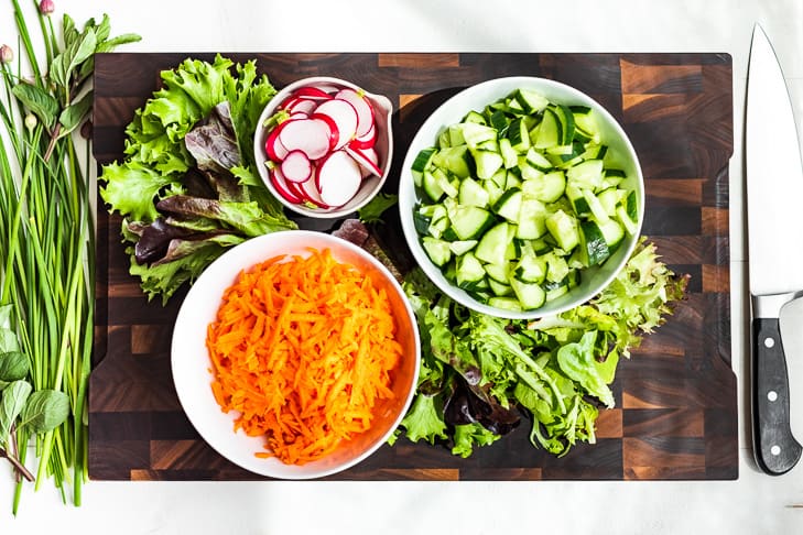 Chopped cucumbers, sliced radishes, pickled carrots, lettuce and fresh herbs on a wooden cutting board.