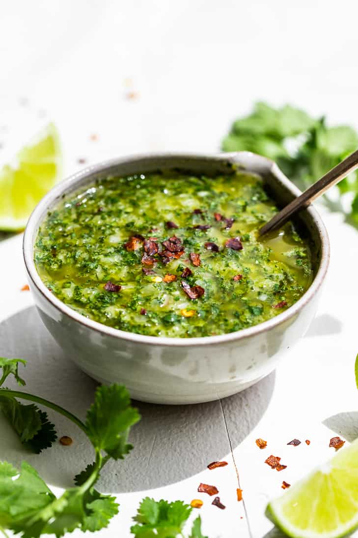 Cilantro Chimichurri in a pottery bowl on a white backdrop with cilantro springs and lime wedges around the bowl.