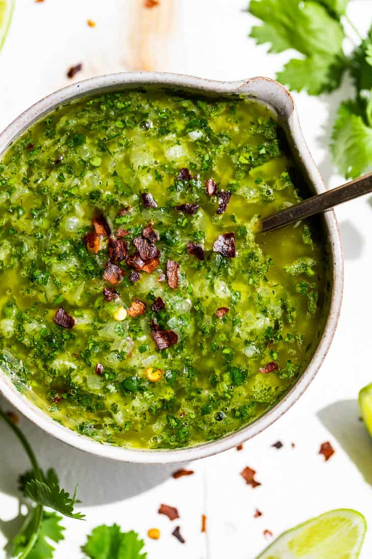 Straight down view of Cilantro Chimichurri in a pottery bowl with a silver spoon with cilantro sprigs and lime wedges around it.