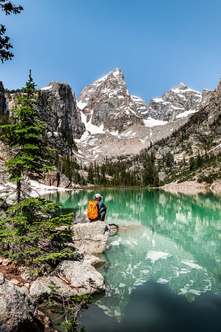 Delta Lake in Grand Teton National Park with a man with an orange backpack sitting on a rock looking at the glacial turquoise lake with snow dotting the mountains behind the lake.