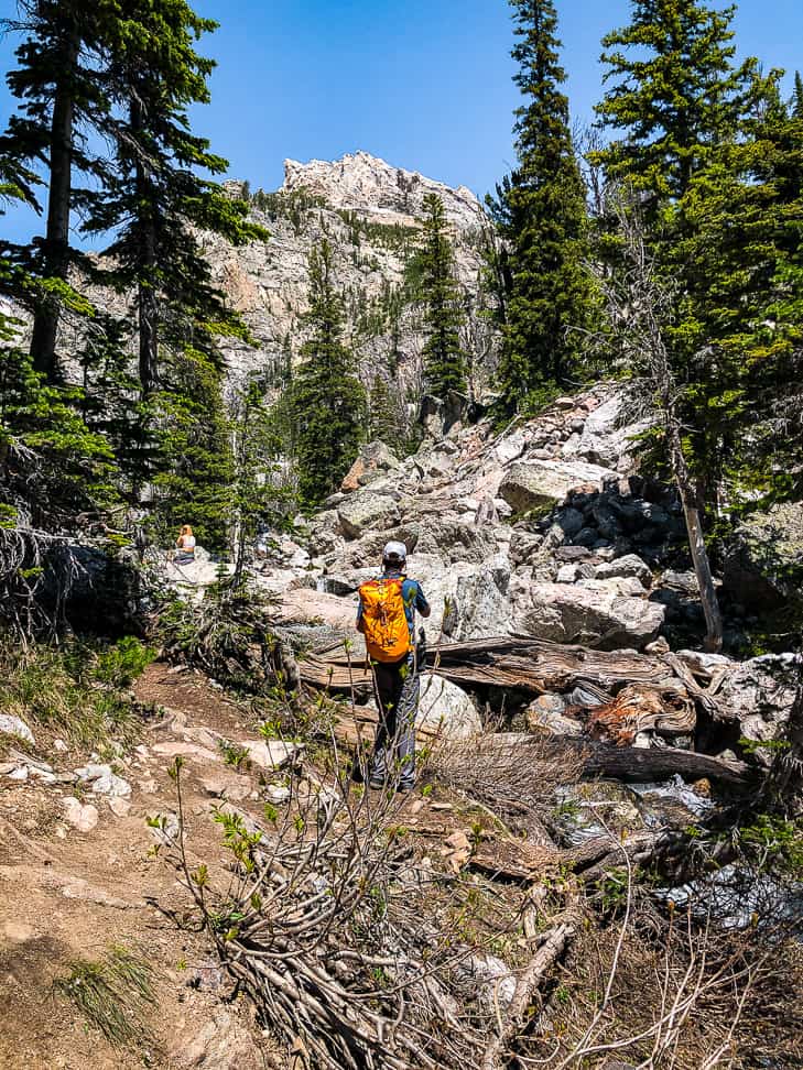 Hiker with an orange backpack standing near the creek running alongside the trail with mountains in the distance.