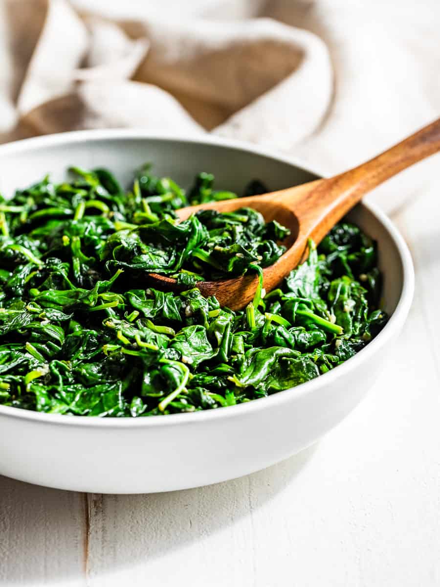 Garlic Sautéed Spinach in a white bowl with a wooden spoon on a white background with a tan linen napkin draped behind it..