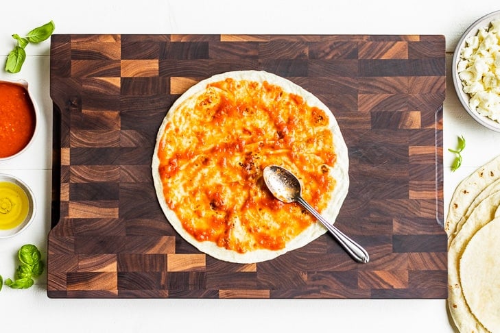 Spreading the pizza sauce over a tortilla on a wooden cutting board.