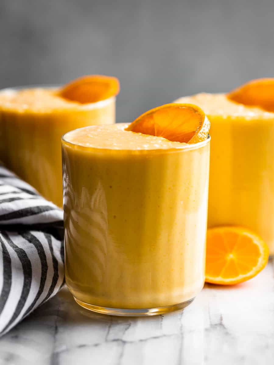 3 glasses of Orange Creamsicle Smoothie on a marble background with a striped linen on the side and orange slices on top of the glasses.
