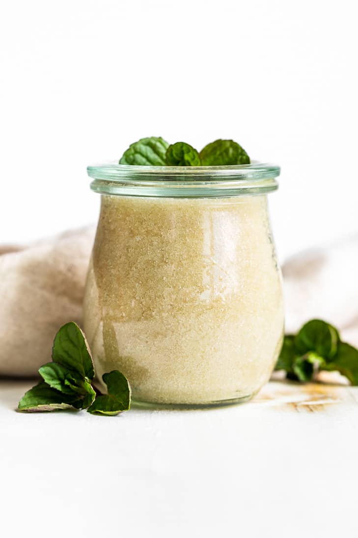 Peppermint Sugar Scrub in a glass jar on a white background with mint springs around it.