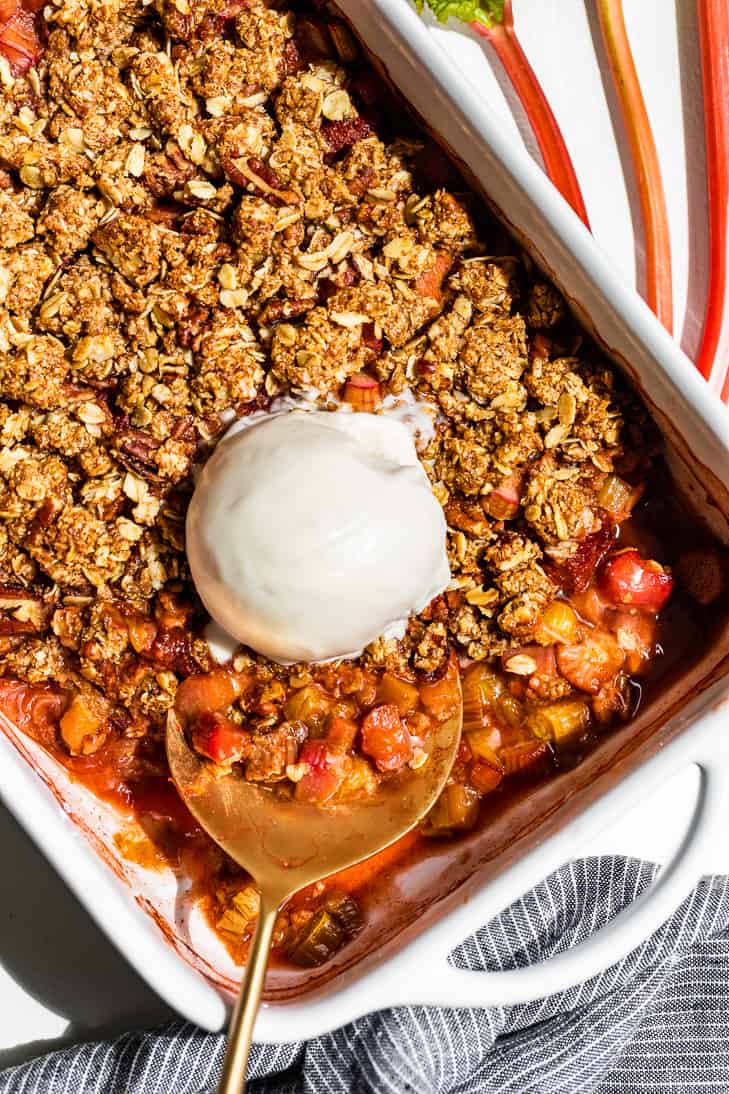 Strawberry Rhubarb Crisp in a white baking dish with a large scoop of ice cream and a gold serving spoon.
