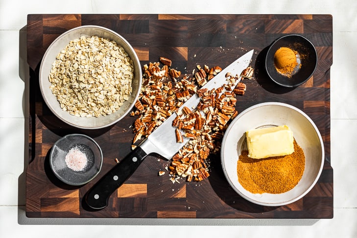 All the ingredients for the crisp topping portioned out on a wooden cutting board and knife chopping up nuts.