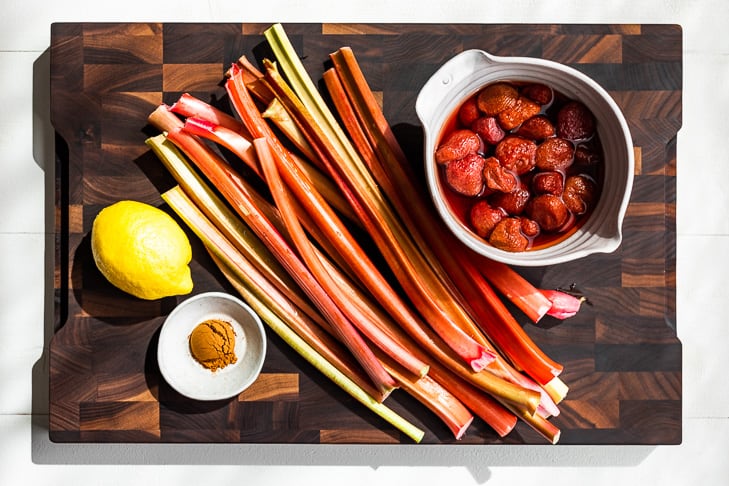Rhubarb stalks on a wooden cutting board with a lemon and a bowl of thawed frozen strawberries and ground cinnamon.