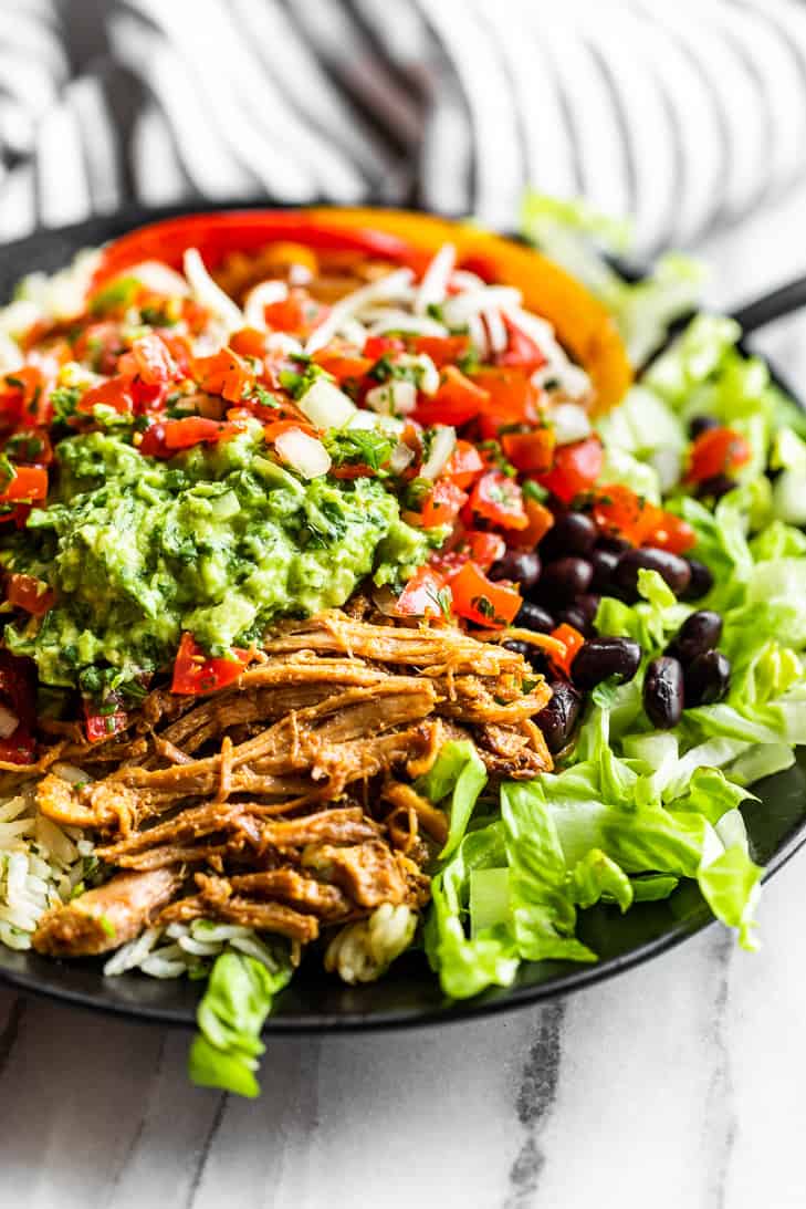 Up close side view of Carnitas Burrito Bowls in a black bowl on a marble background with a black and white striped linen on the side.