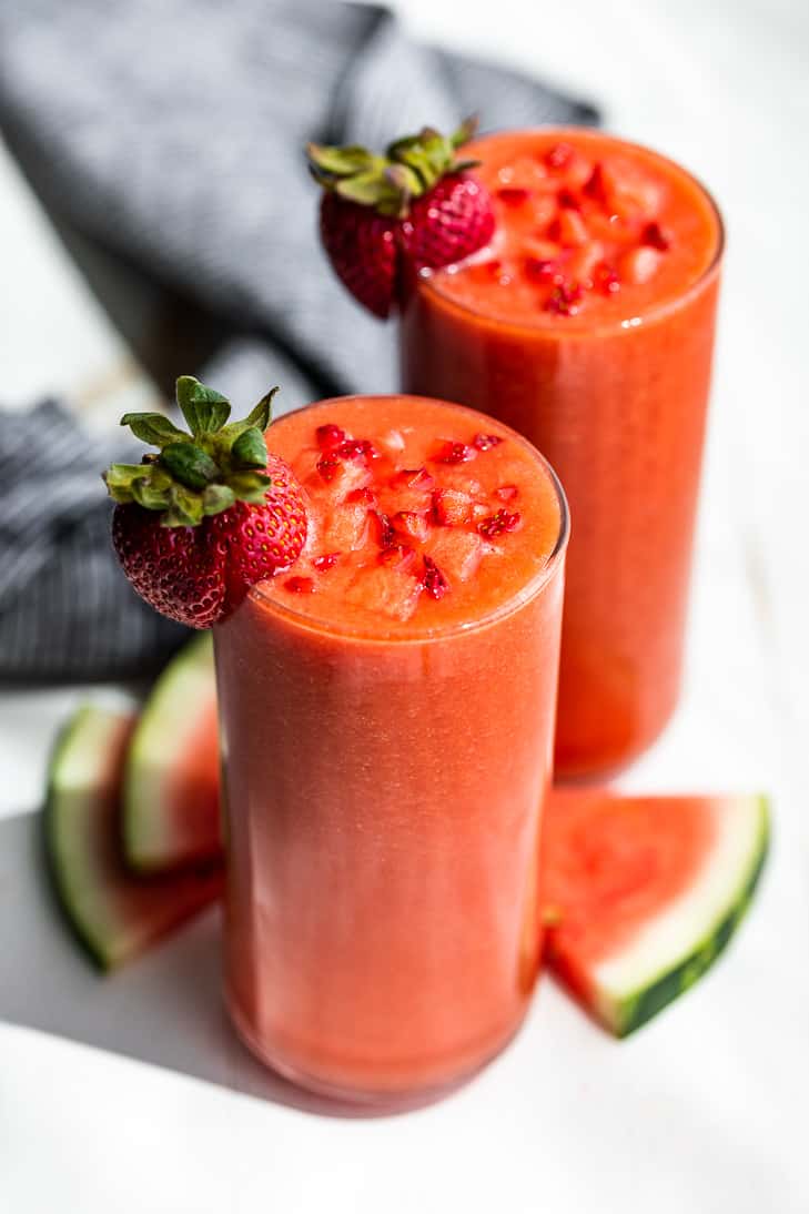 A side view of 2 glasses Strawberry Watermelon Smoothie with watermelon cubes on top, watermelon slices alongside, and 2 strawberries on the rims of the glasses.