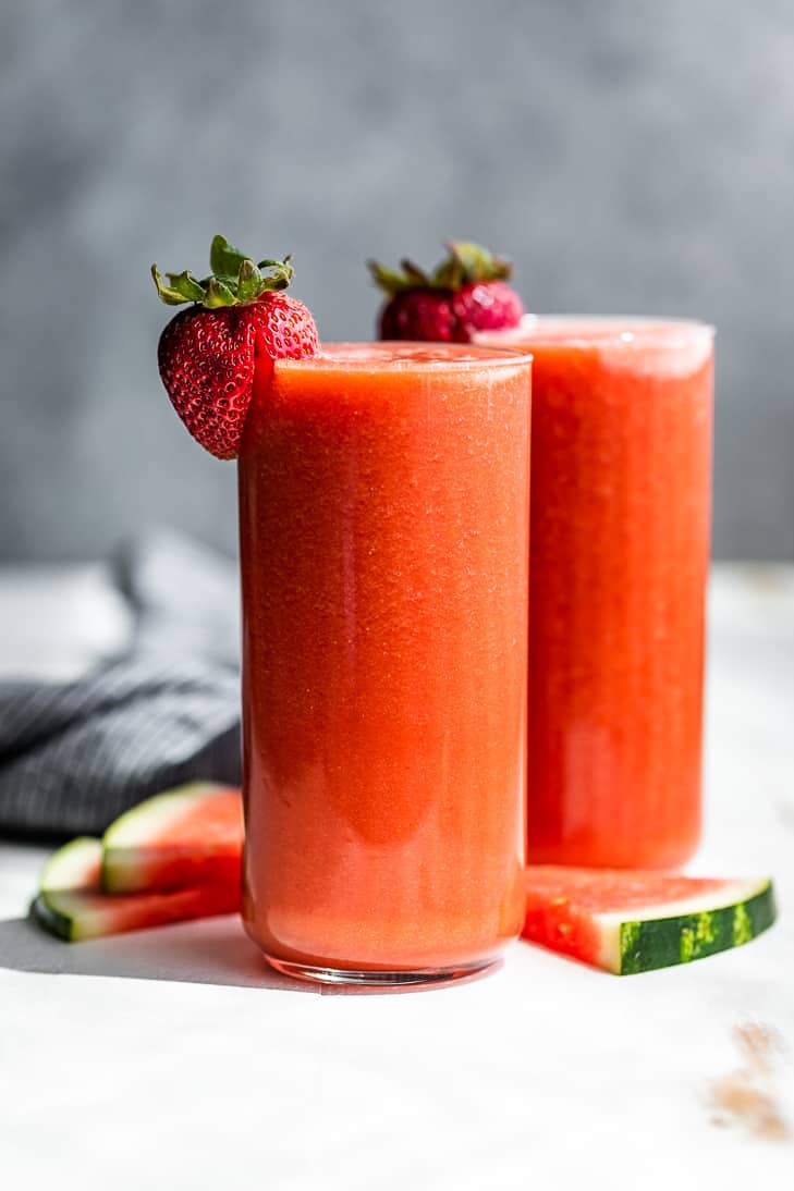 Two tall glasses of Strawberry Watermelon Smoothie against a grey backdrop with watermelon slices around them and a strawberry on the sides of the glasses.
